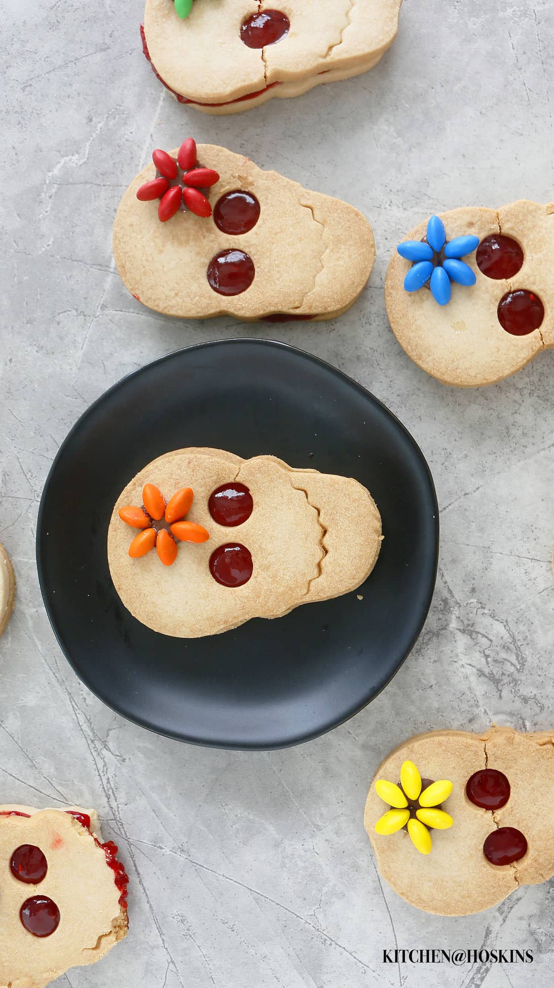 Cute Halloween skull cookies decorated with raspberry jam and M&M flower, all displayed on a board with one on a black plate