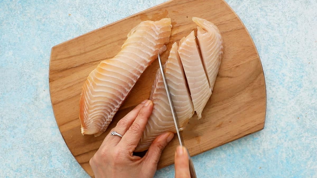 two hands cutting one tilapia fish fillet into sticks.