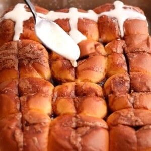 a hand pouring white icing over brown baked rolls.