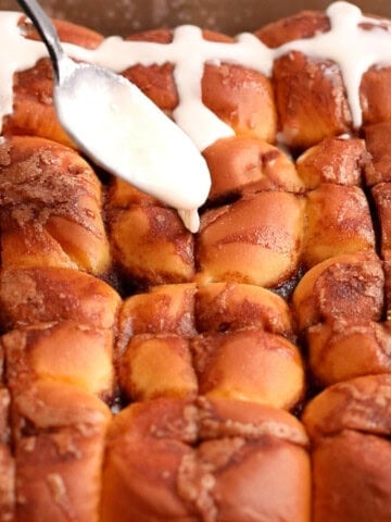 a hand pouring white icing over brown baked rolls.
