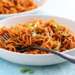 red colored bbq ramen noodles in a white bowl along with a black fork.