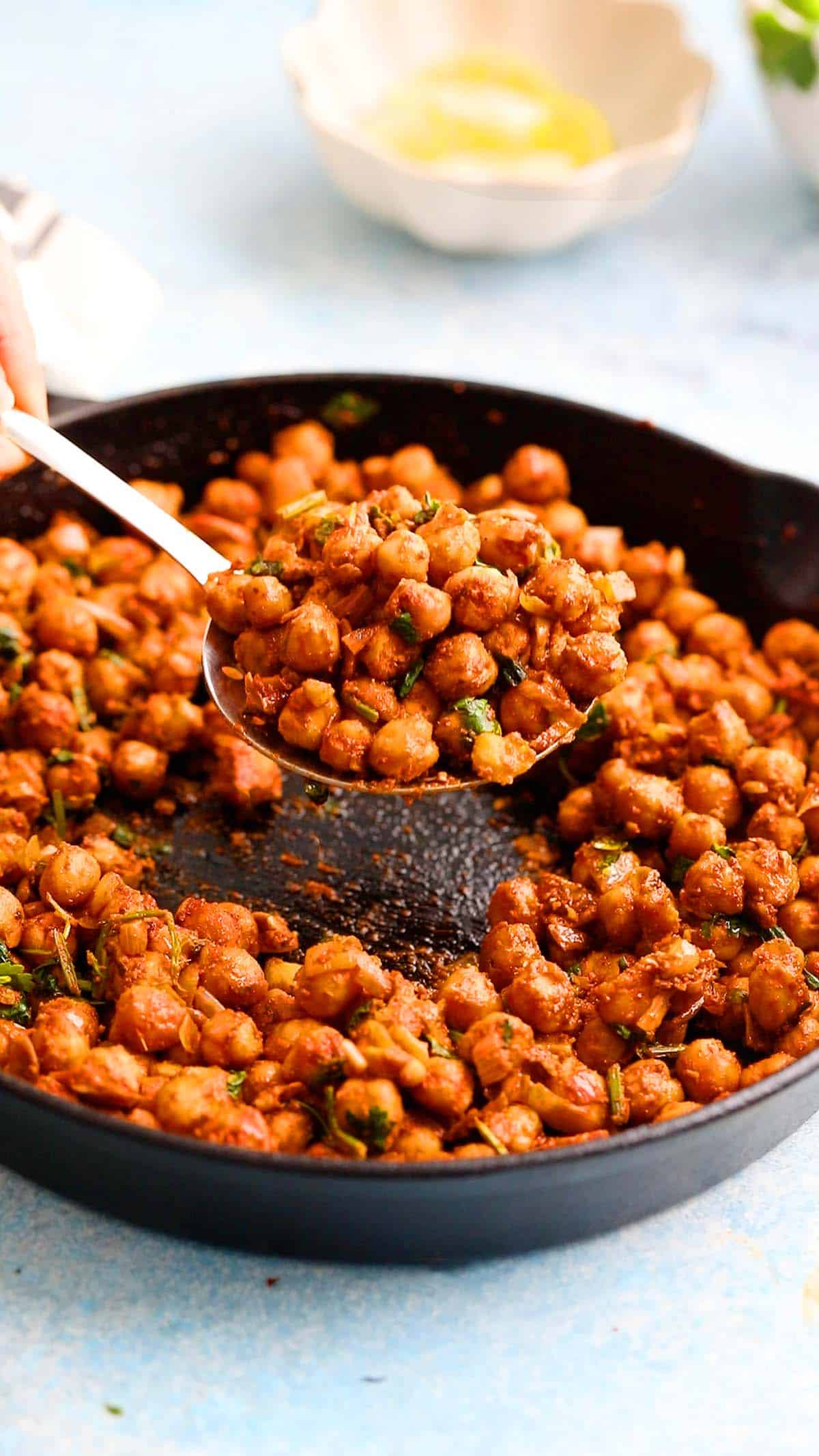 a hand lifting cooked spicy chickpeas from a black skillet using a big spoon. 