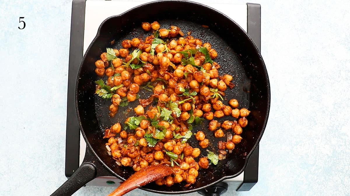 a hand sauteing chickpeas and spices in a black skillet using a wooden spoon.