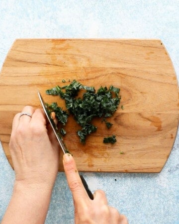 two hands chopping kale leaf on a wooden board.