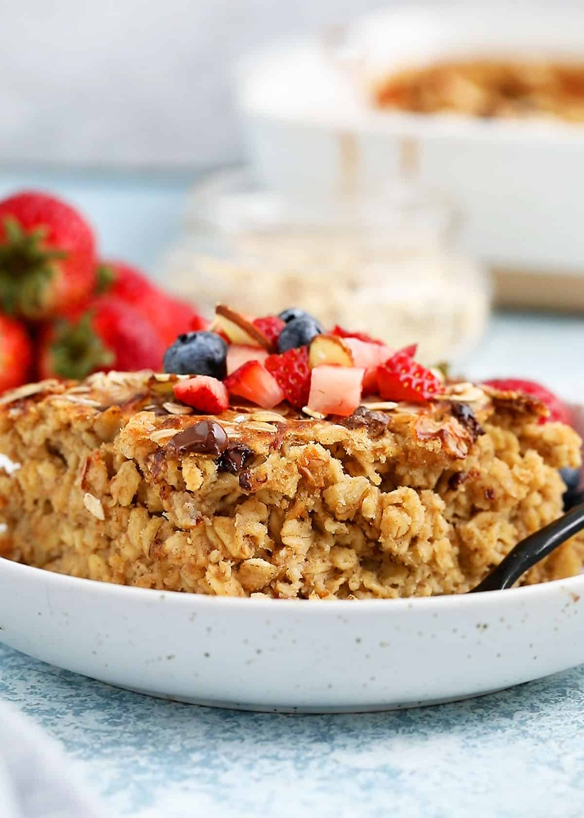 one square piece of baked oatmeal topped with berries in a white plate along with a black spoon.