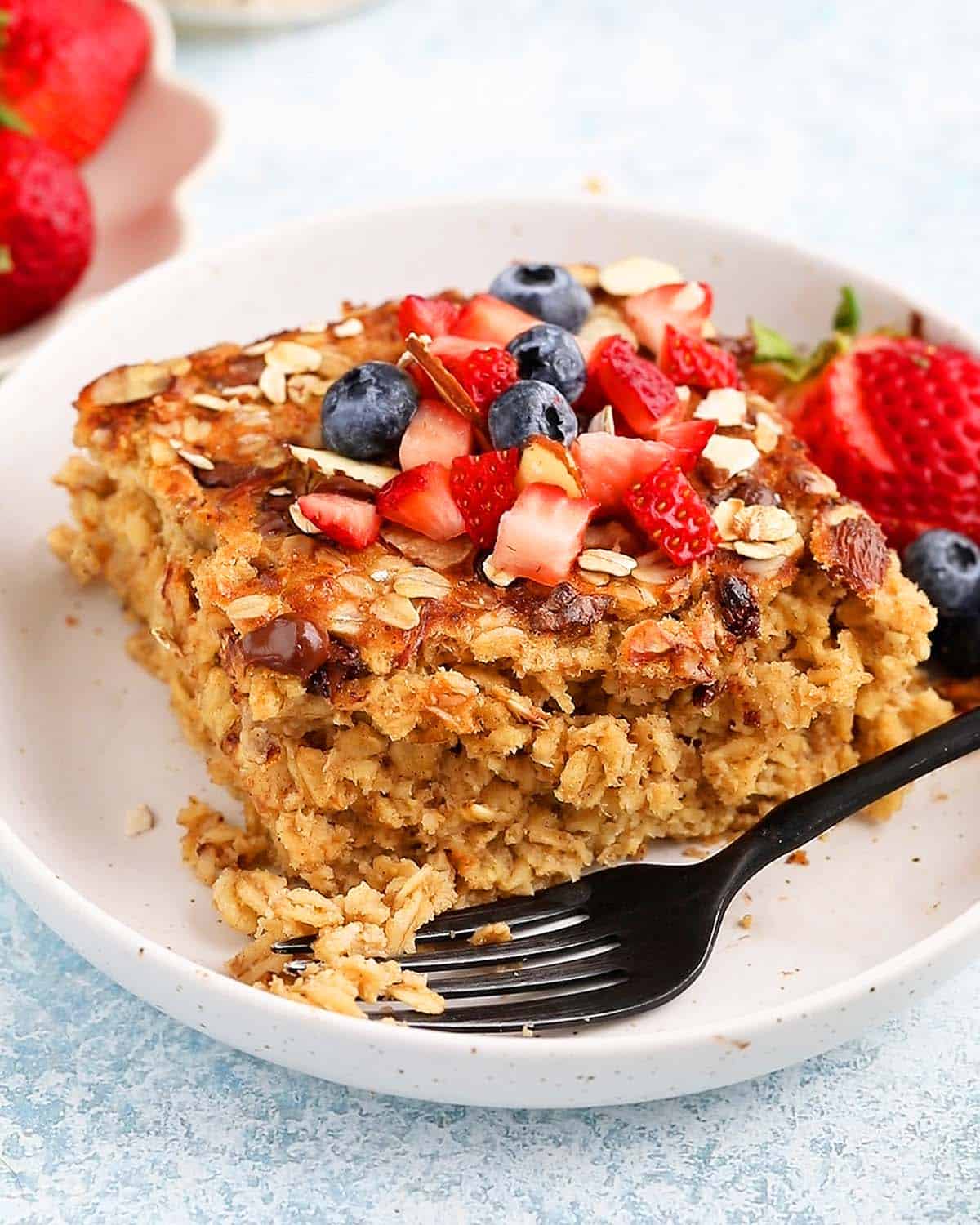 one square piece of baked oatmeal topped with berries in a white plate along with a black spoon.