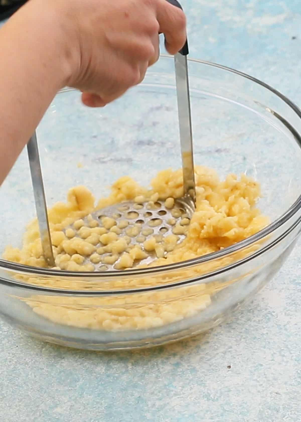 a hand mashing bananas in a glass bowl using a potato masher.