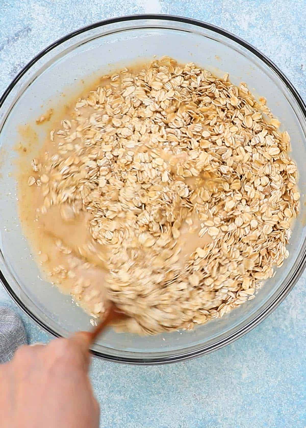 a hand stirring oats along with wet ingredients in a glass bowl.