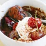 White mug with chocolate cake, topped with whipped cream and 2 fresh cherries.