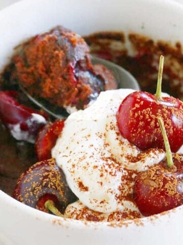 White mug with chocolate cake, topped with whipped cream and 2 fresh cherries.