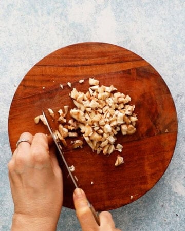 two hands chopping mushrooms into small pieces on a round wooden cutting board.