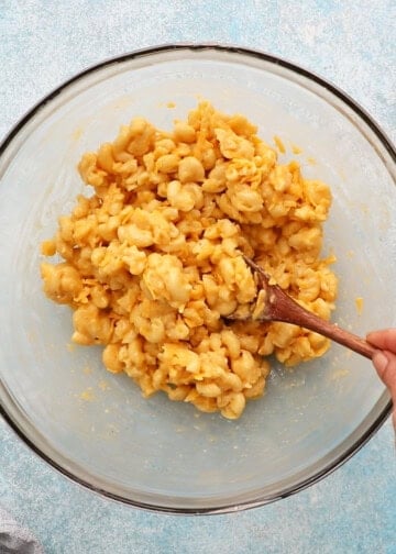 a hand mixing cooked macaroni and cheese in a glass bowl.