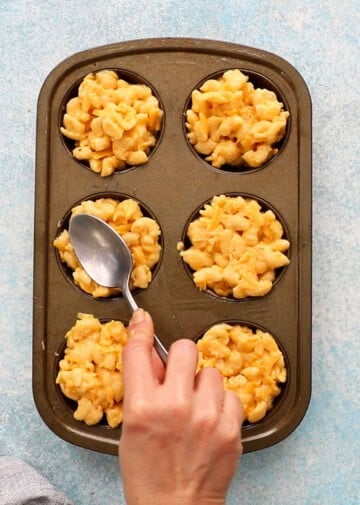 a hand adding yelllow macaroni and cheese into a metal muffin pan using a spoon.