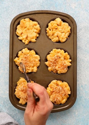 a hand adding brown breadcrumbs  over mac and cheese in a metal muffin pan.
