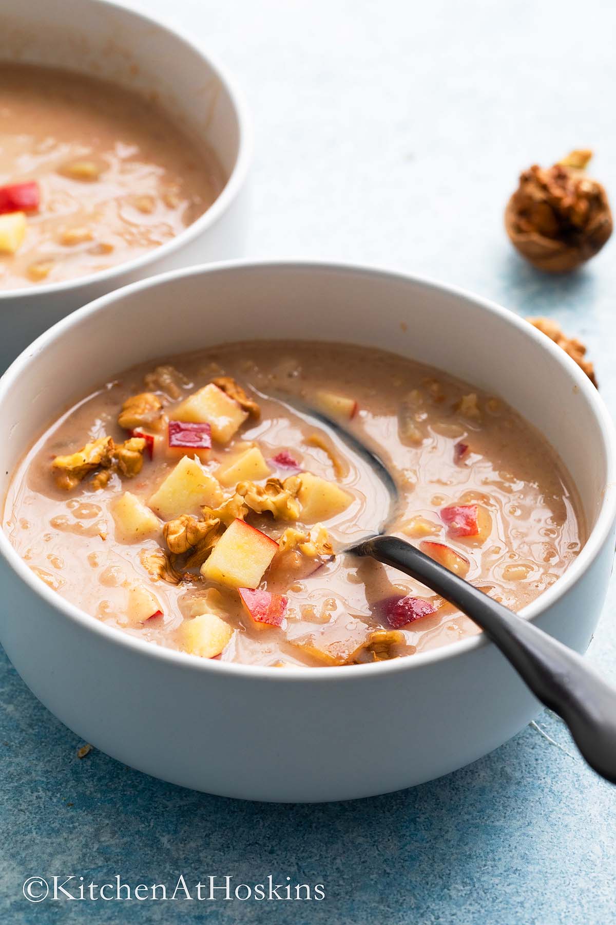 2 white bowls with oatmeal topped with chopped apples, walnuts and a black spoon.