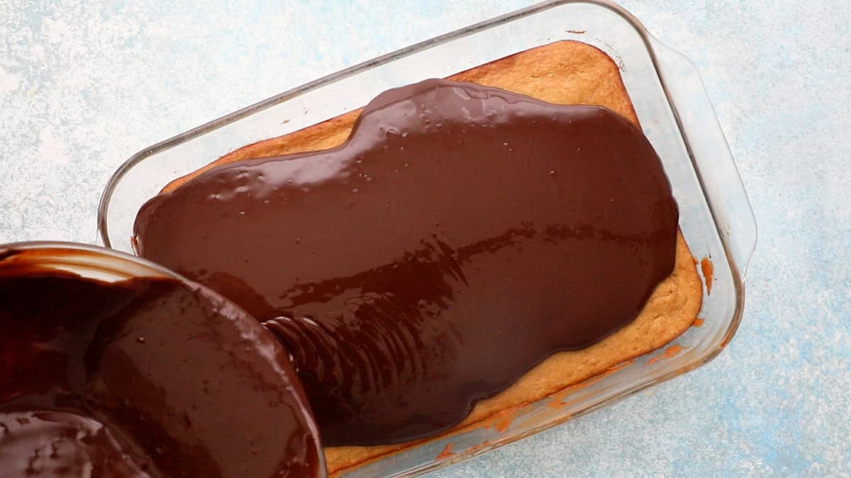 a hand pouring brown chocolate glaze over baked banana sheet cake.