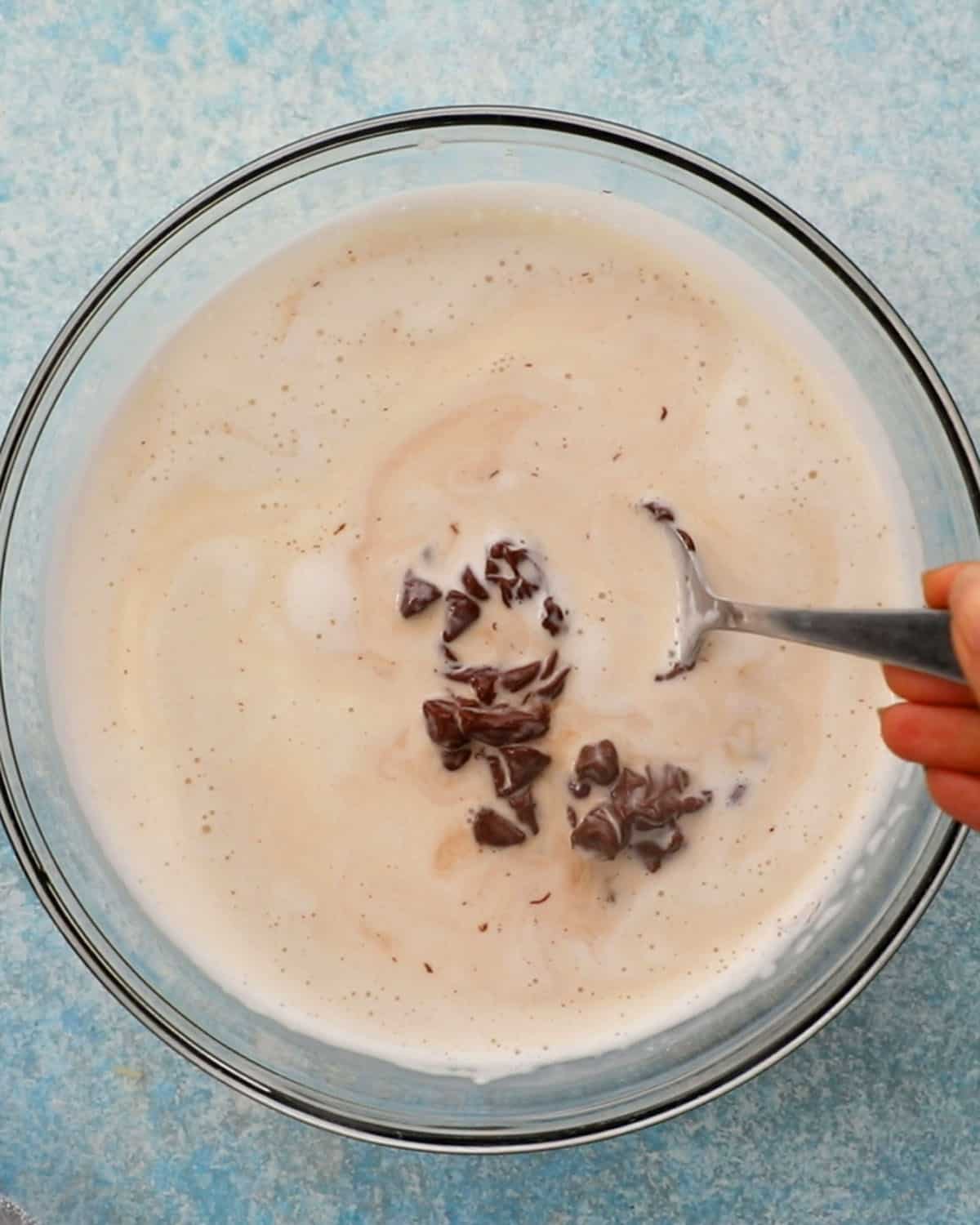 a hand mixing white cream and brown chocolate in a glass bowl.