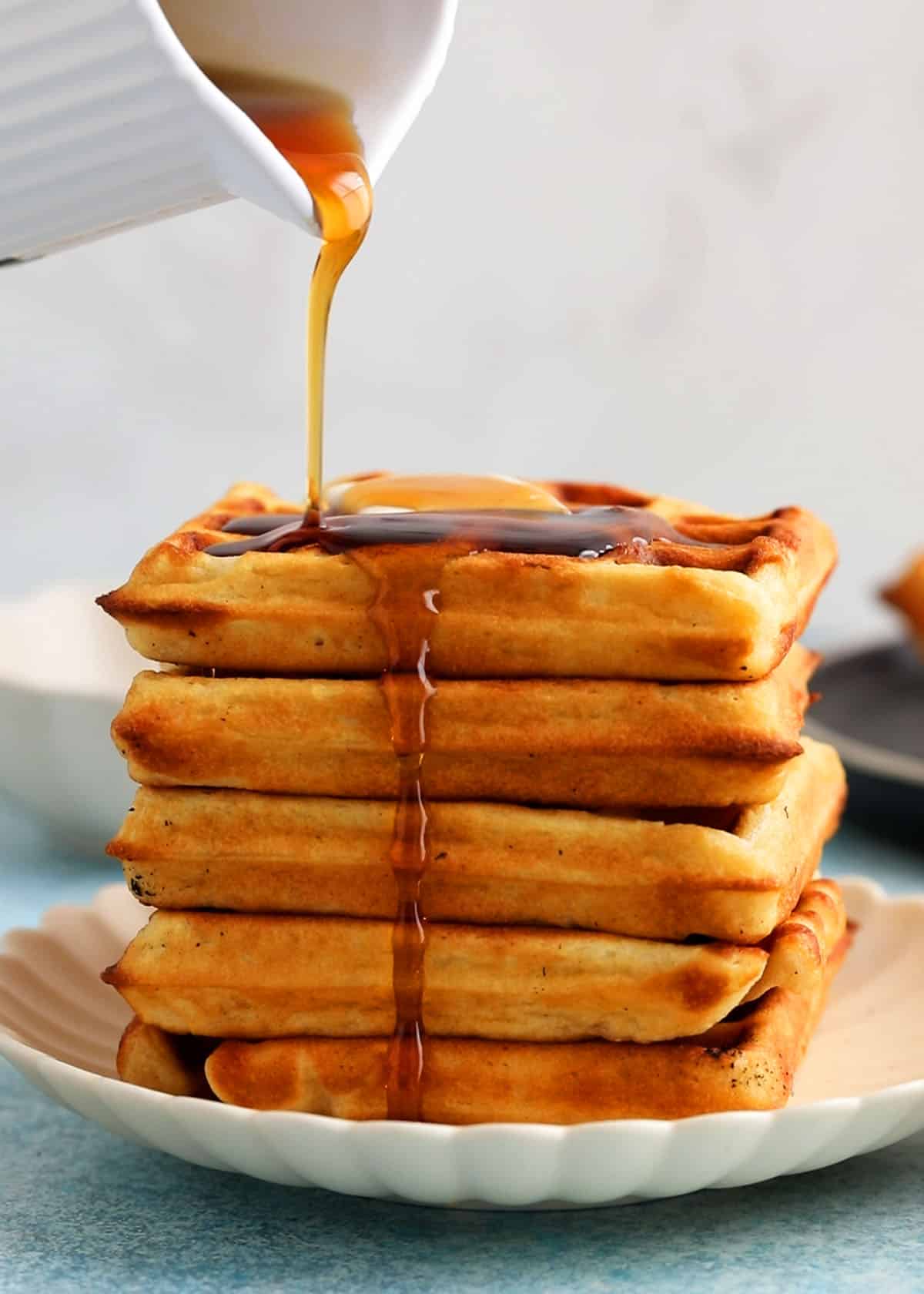 5 waffles stacked on a white plate and topped with maple syrup.