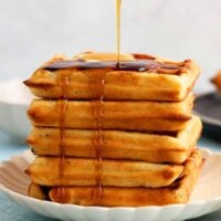 5 waffles stacked on a white plate and topped with maple syrup.