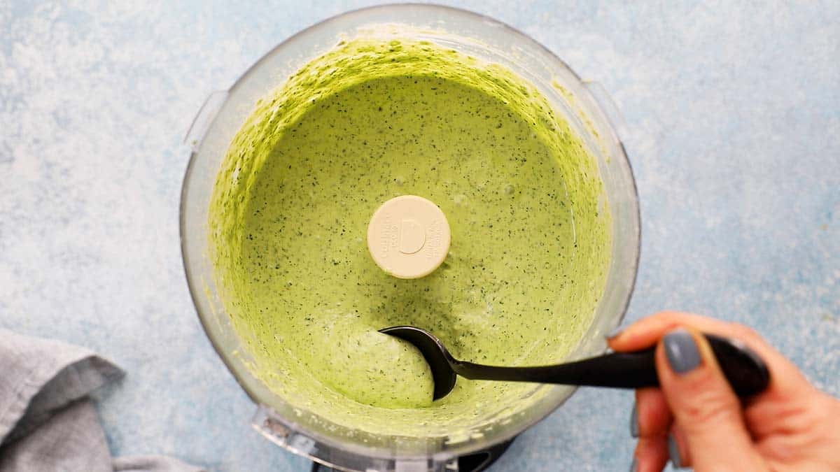 removing some green sauce with a black spoon from a food processor.