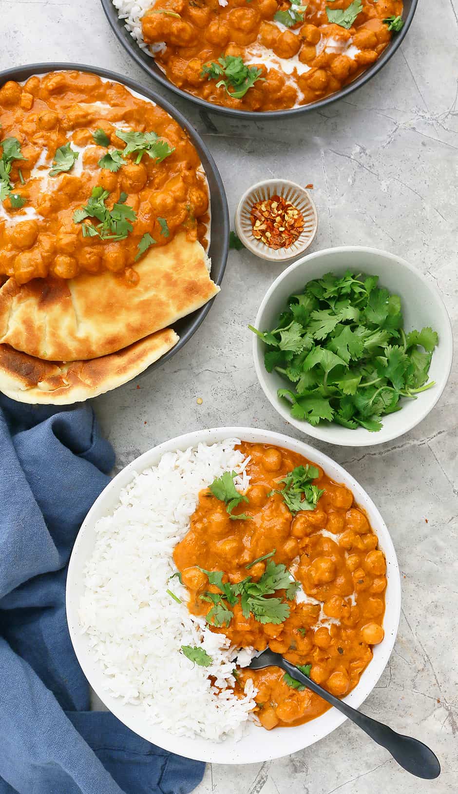 bowls of creamy Indian chickpeas curry with white rice and naan, a small bowl with fresh cilantro and a tiny bowl of red pepper flakes
