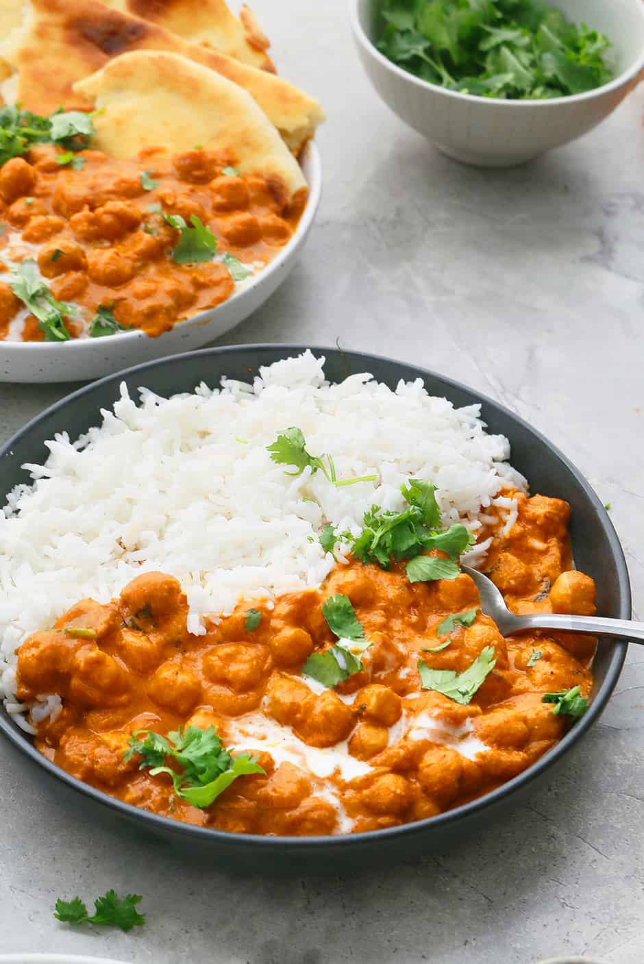 a black bowl filled with butter chickpeas curry and white rice with a spoon. Another white bowl with the same chickpeas and naan instead