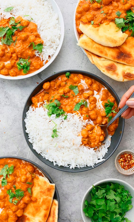 white and black bowls filled with butter chickpeas, white rice and naan, garnished with cilantro and a hand trying to take a bite from one of teh black bowls.