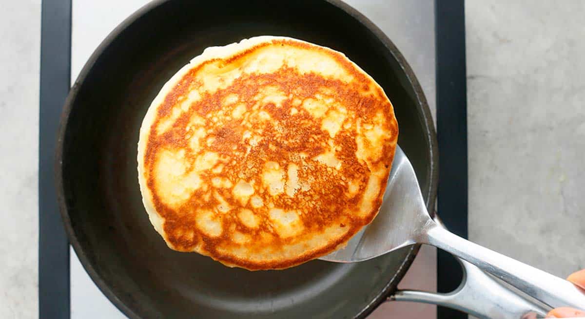 a flat stainless steel spatula with one pancake above a black skillet.
