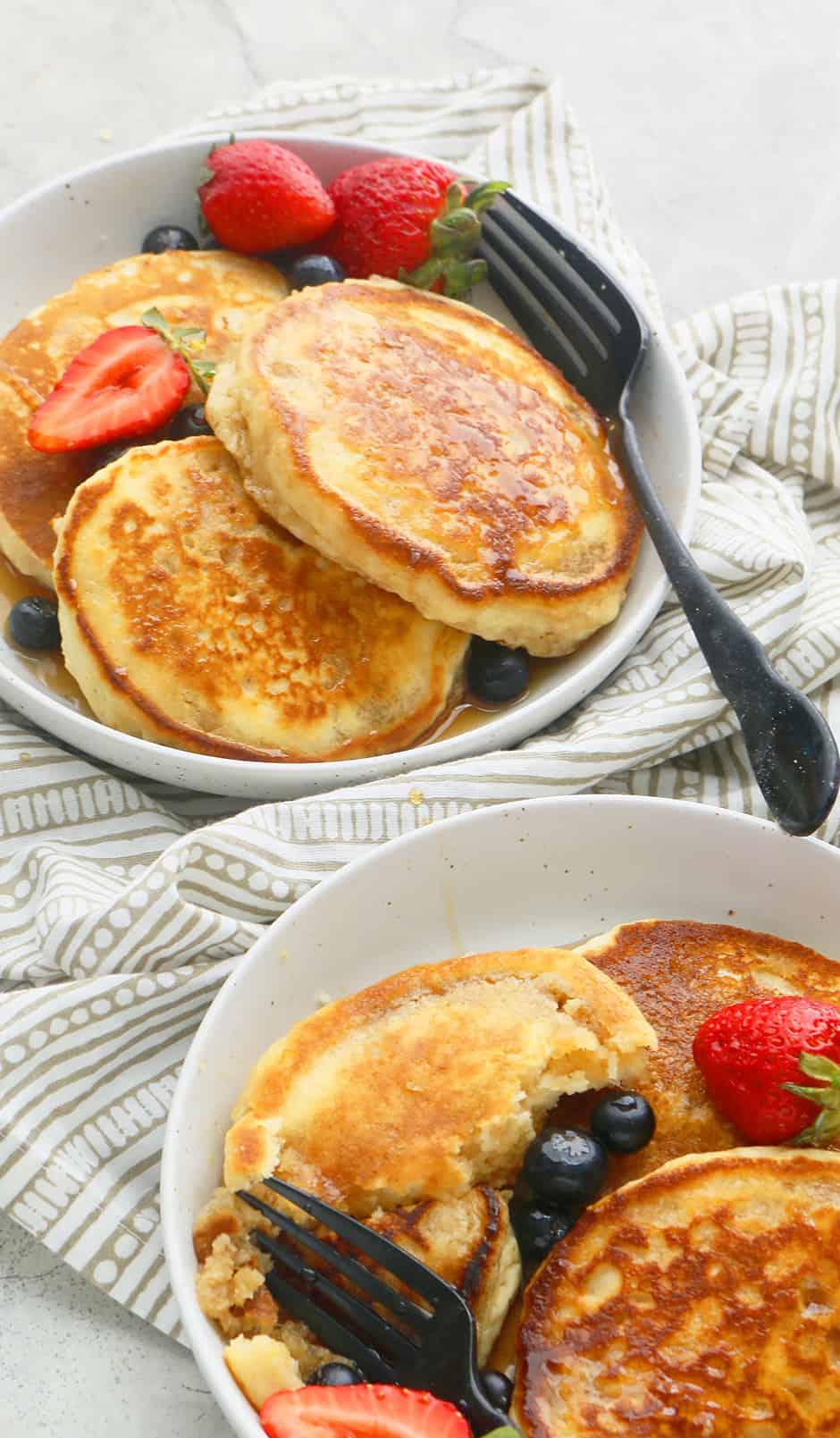 2 white plates each filled with eggless pancakes, topped with maple syrup, garnished with fresh berries  and with black forks on the side