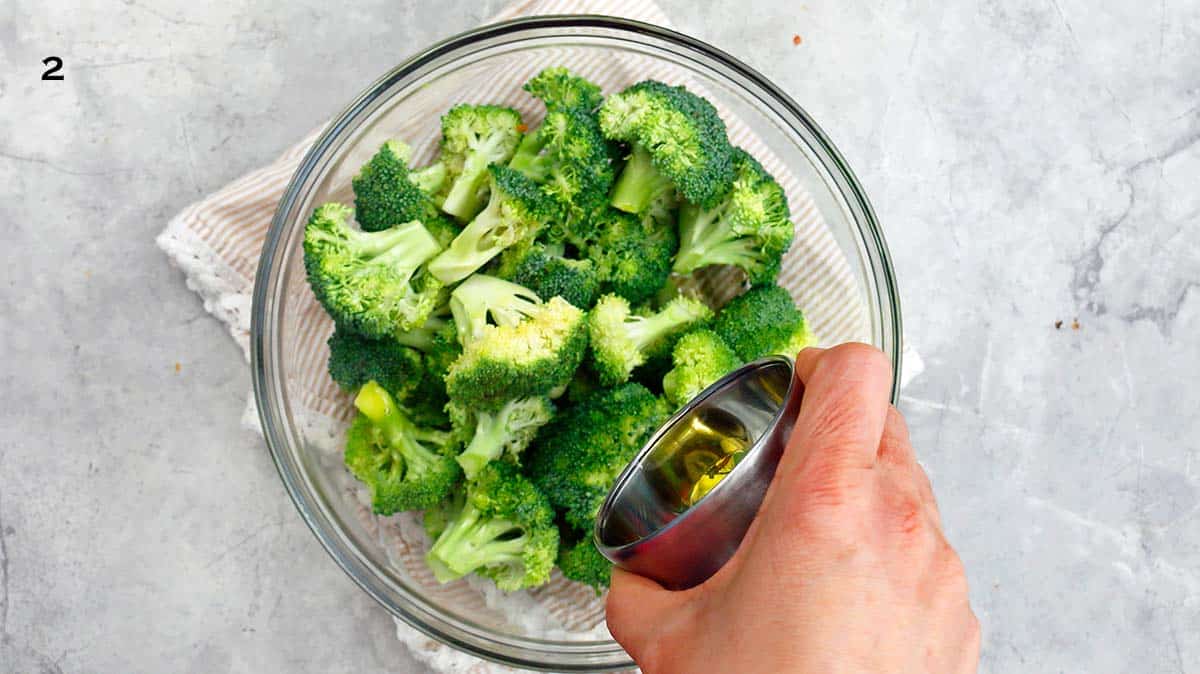 a hand pouring oil into a glass bowl with broccoli florets.