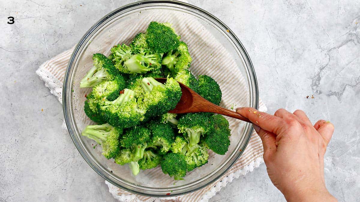 A hand tossing broccoli florets with a wooden spoon.