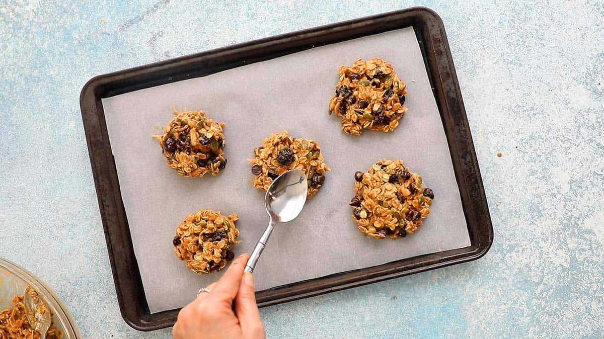 a hand flattening cookie dough using a spoon on a baking sheet,