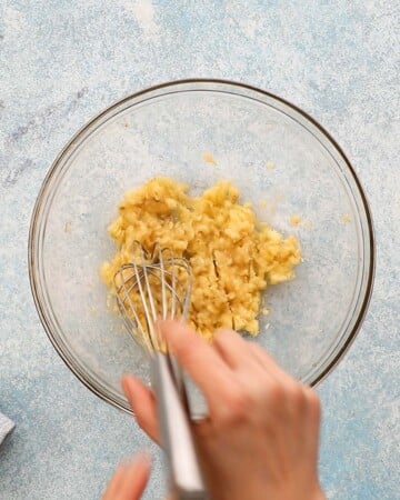 a hand mashing bananas in a glass bowl using a metal whisk.