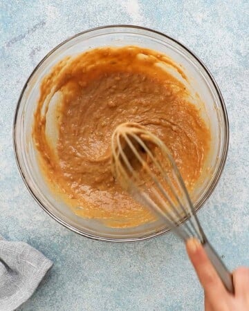 two hands mixing brown wet ingredients in a glass bowl.