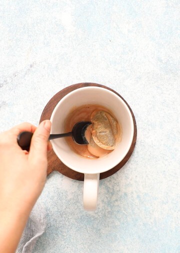 a hand stirring tea bags in a white mug with a black spoon.