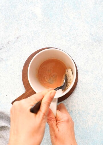 a hand stirring tea bags in a white mug with a black spoon.
