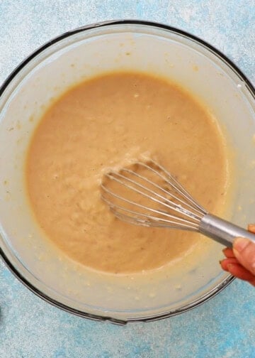 a hand whisking brown banana bread batter in a large glass bowl.