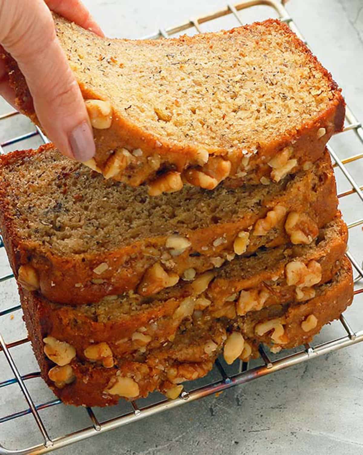 a hand removing holding the first of 4 stacked slices of banana bread placed on a wire rack.