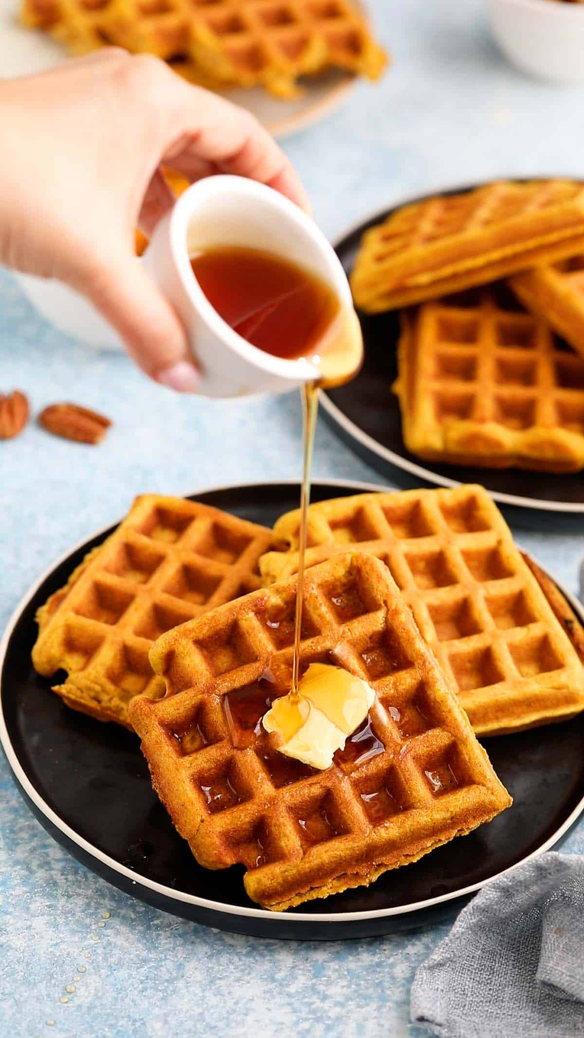 a hand pouring syrup on top of 3 waffles placed on a black plate, topped with some butter.