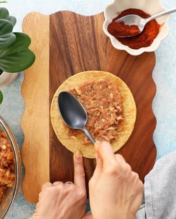 a hand spreading ground chicken on a yellow corn tortilla using a black spoon.