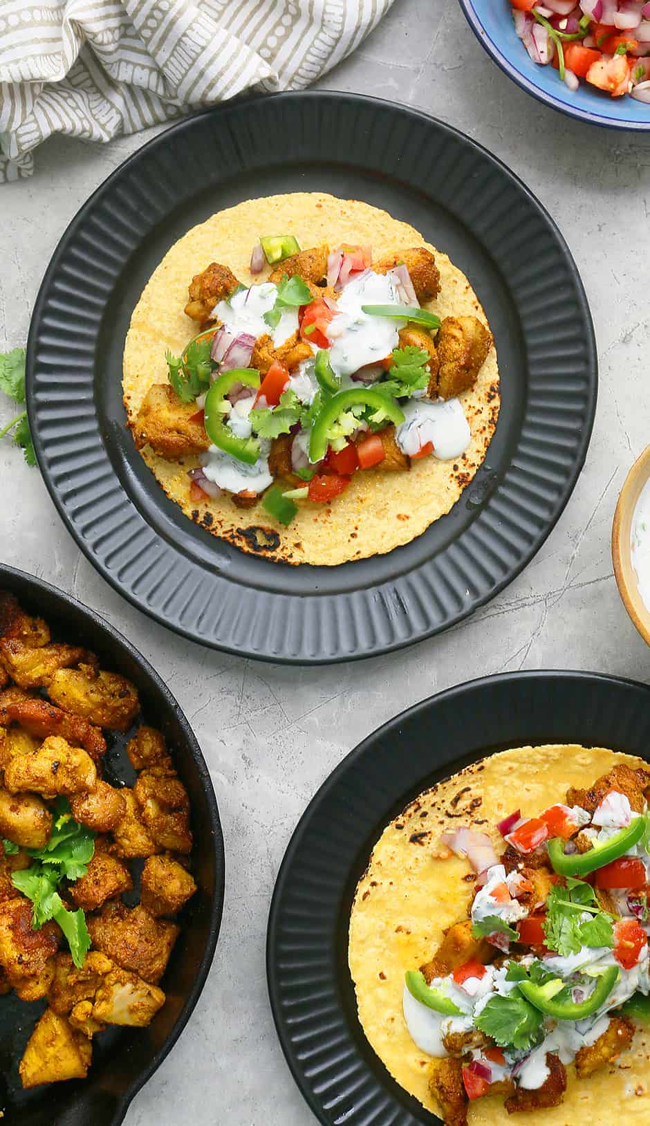 2 black plates with spicy chicken tacos, a blue bowl with pico de gallo and more spicy chicken