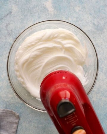 a hand beating white egg whites in a large glass bowl using a red beater.