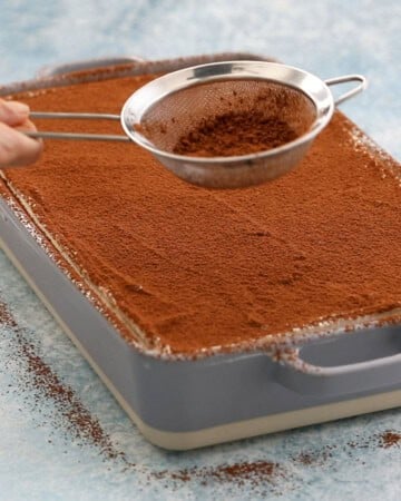 a hand sprinkling brown cocoa powder over a baked tres leches cak in a rectangular grey pan.