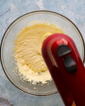 a hand beating yellow egg yolks in a large glass bowl using a red beater.