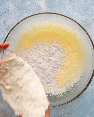 a hand adding white flour into a glass bowl.