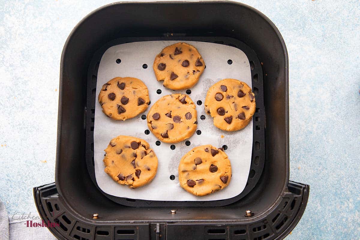 6 shaped chocolate chip cookie dough on a square parchment in air fryer basket.
