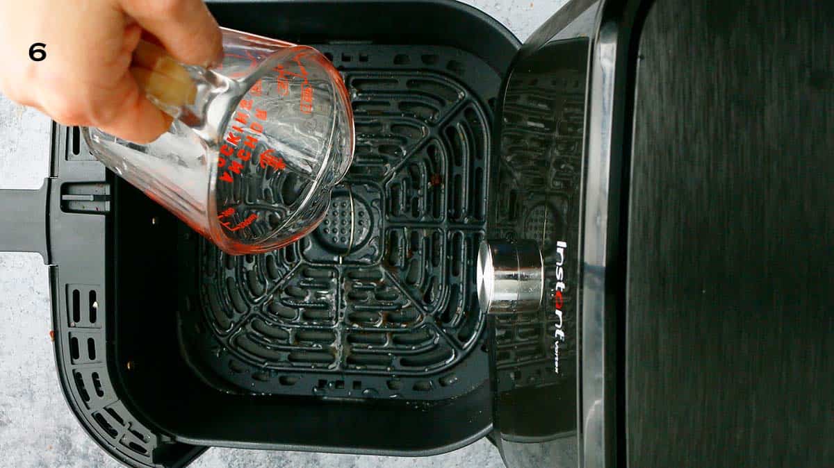 a hand pouring water into a air fryer basket using a glass cup.