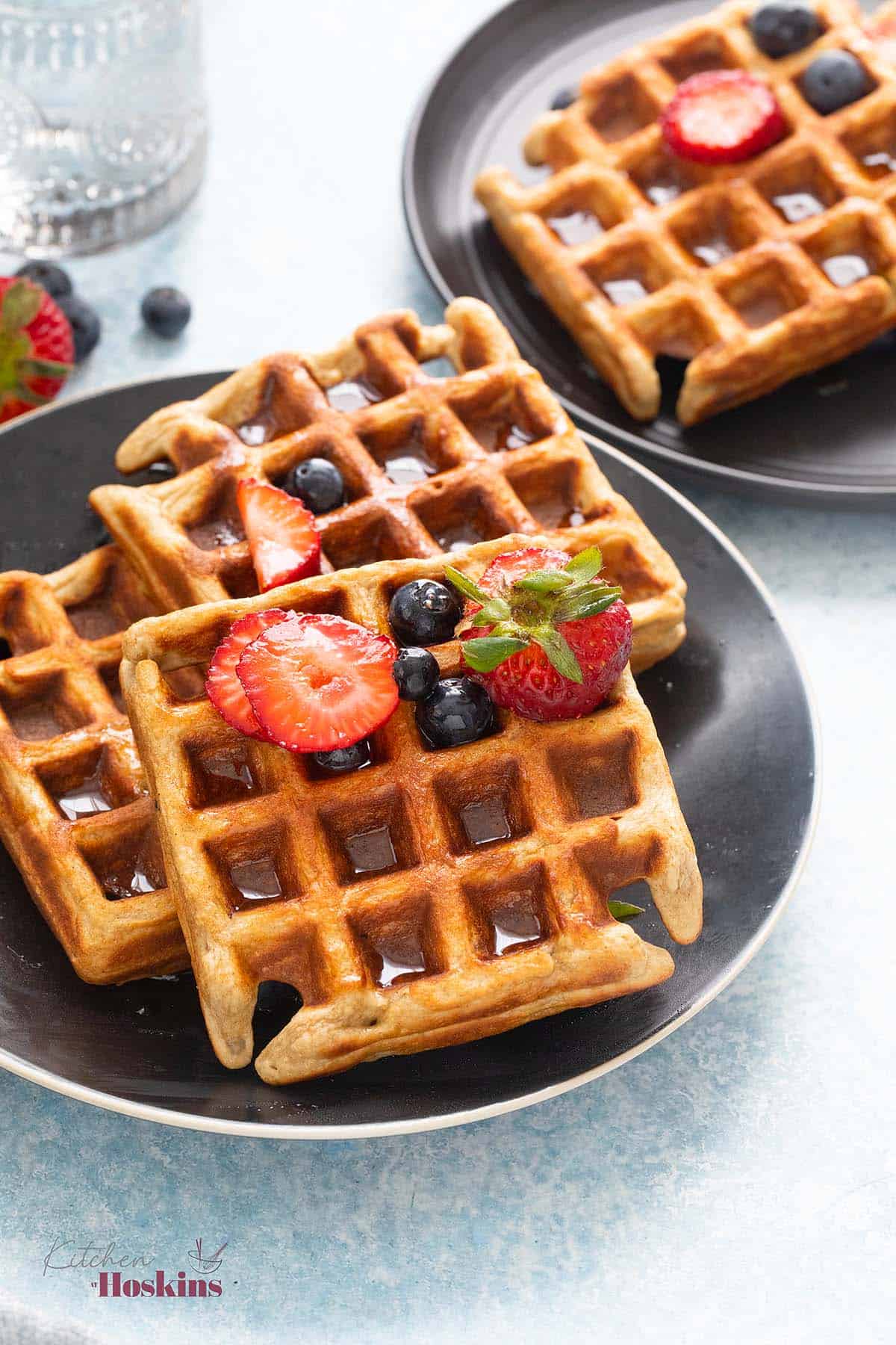 2 black plates with waffles topped with berries and maple syrup.