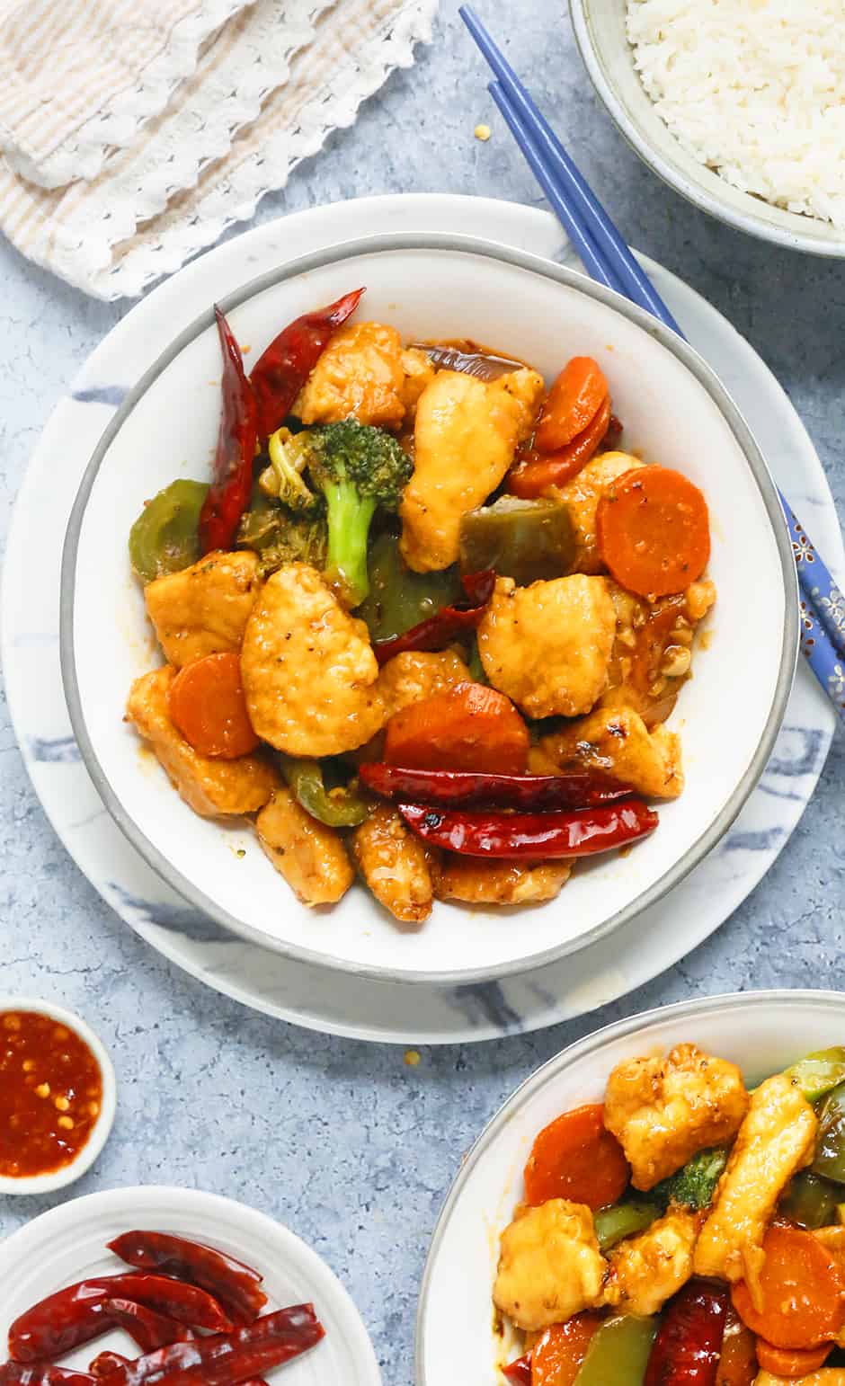 Hunan chicken stir fry in white bowls along with purple chopsticks and red chilies in white bowls on the side