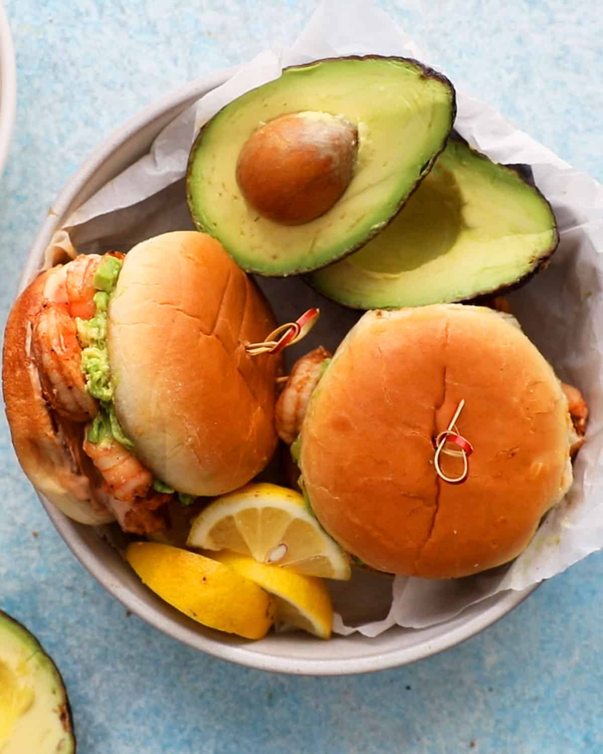 two assembled shrimp sandwich along with one avocado half in a white dish.
